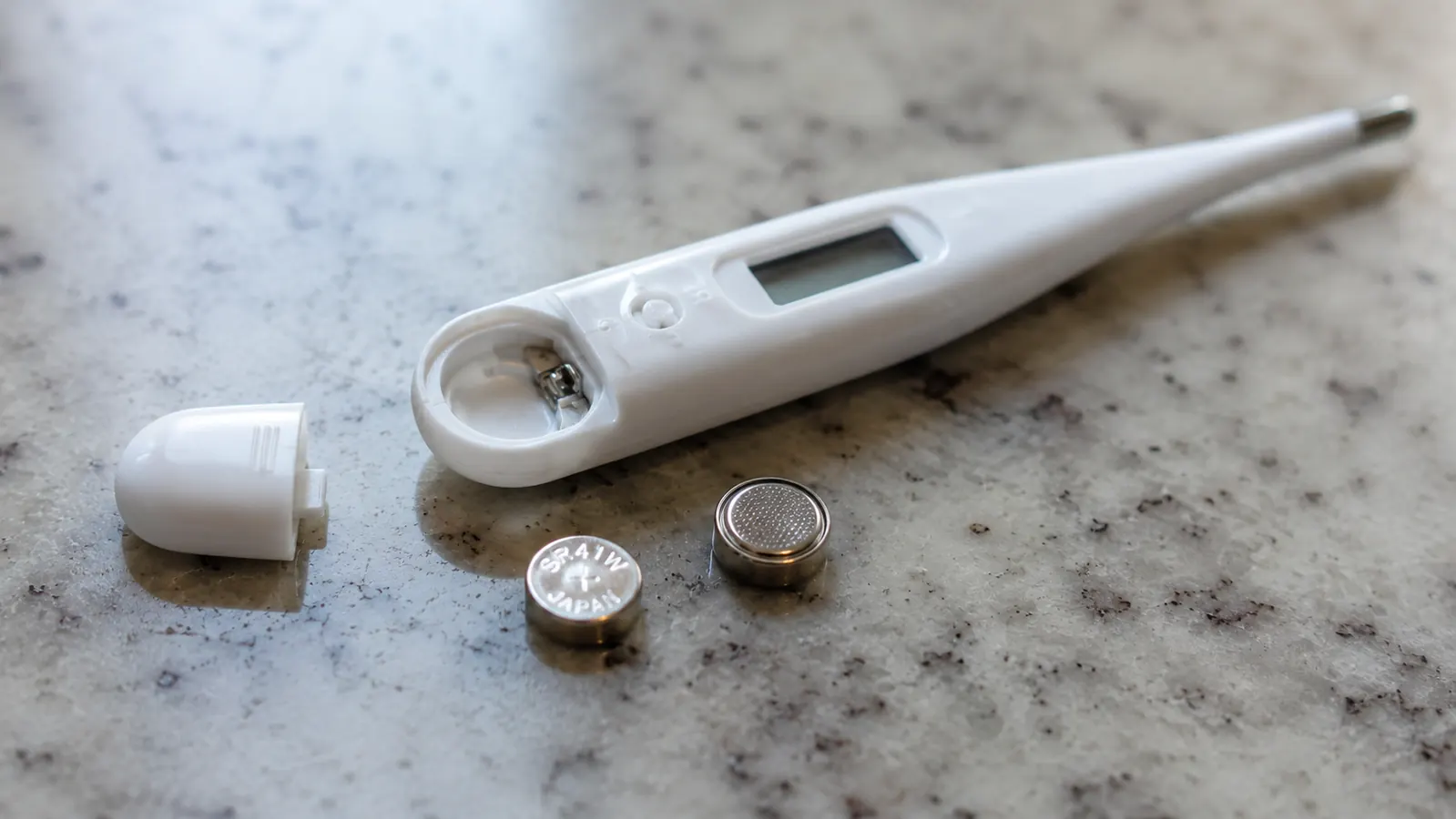 A small button battery and a coin next to the open battery compartment on the back of the thermometer